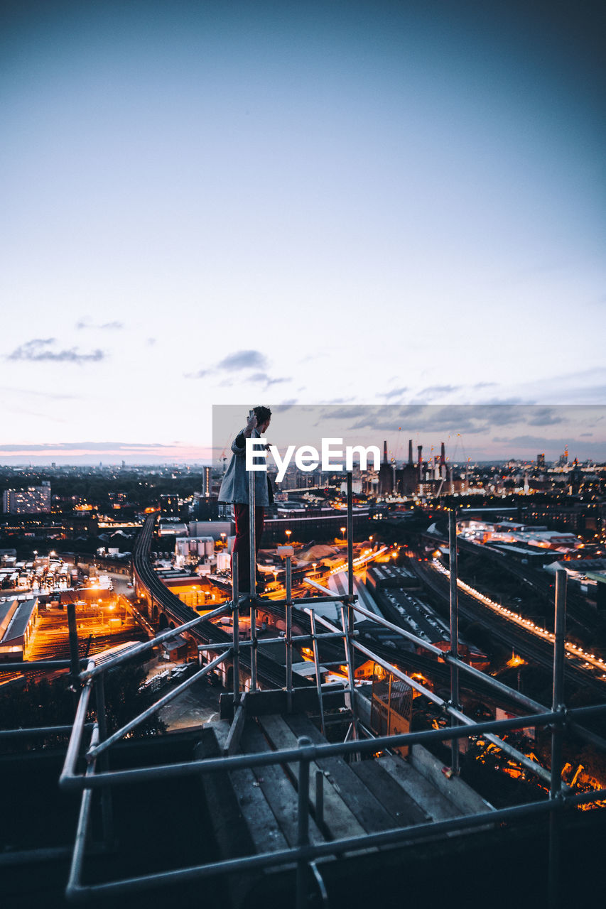 Side view of man standing on built structure against illuminated cityscape during sunset