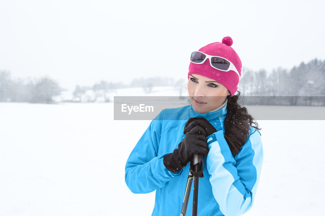 Thoughtful woman holding ski poles while standing on snow covered field
