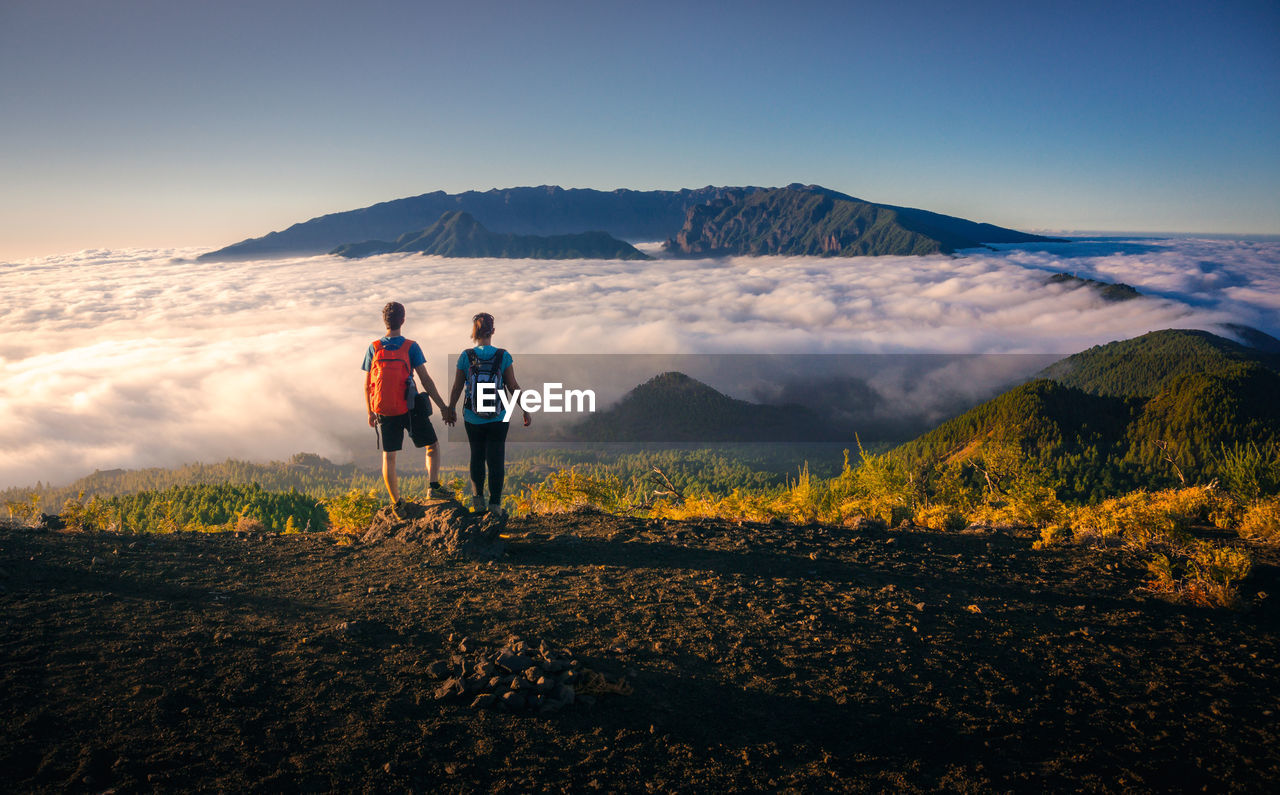 Back view of tender traveling couple standing on hill holding hands while admiring spectacular scenery on clouds over mountainous terrain