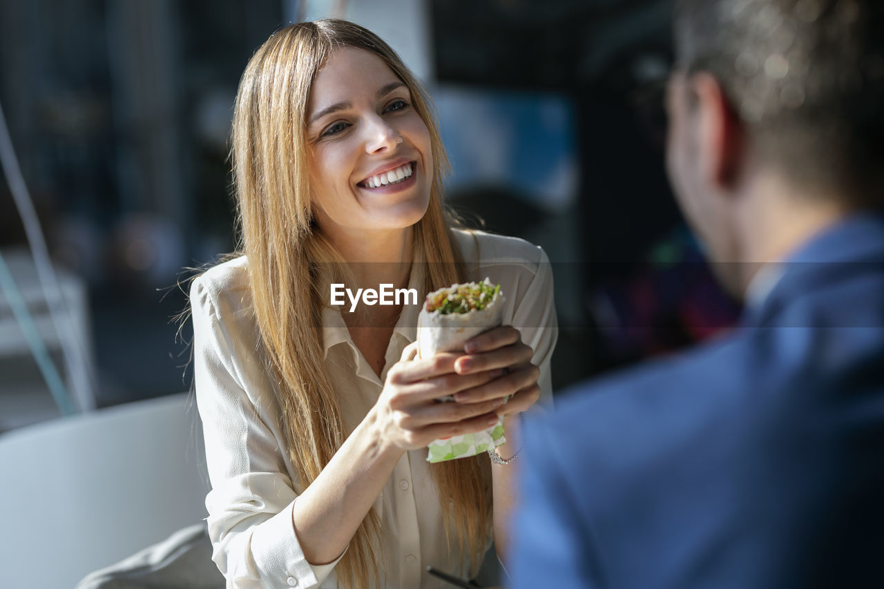 Businesswoman holding food while looking at colleague sitting in cafeteria