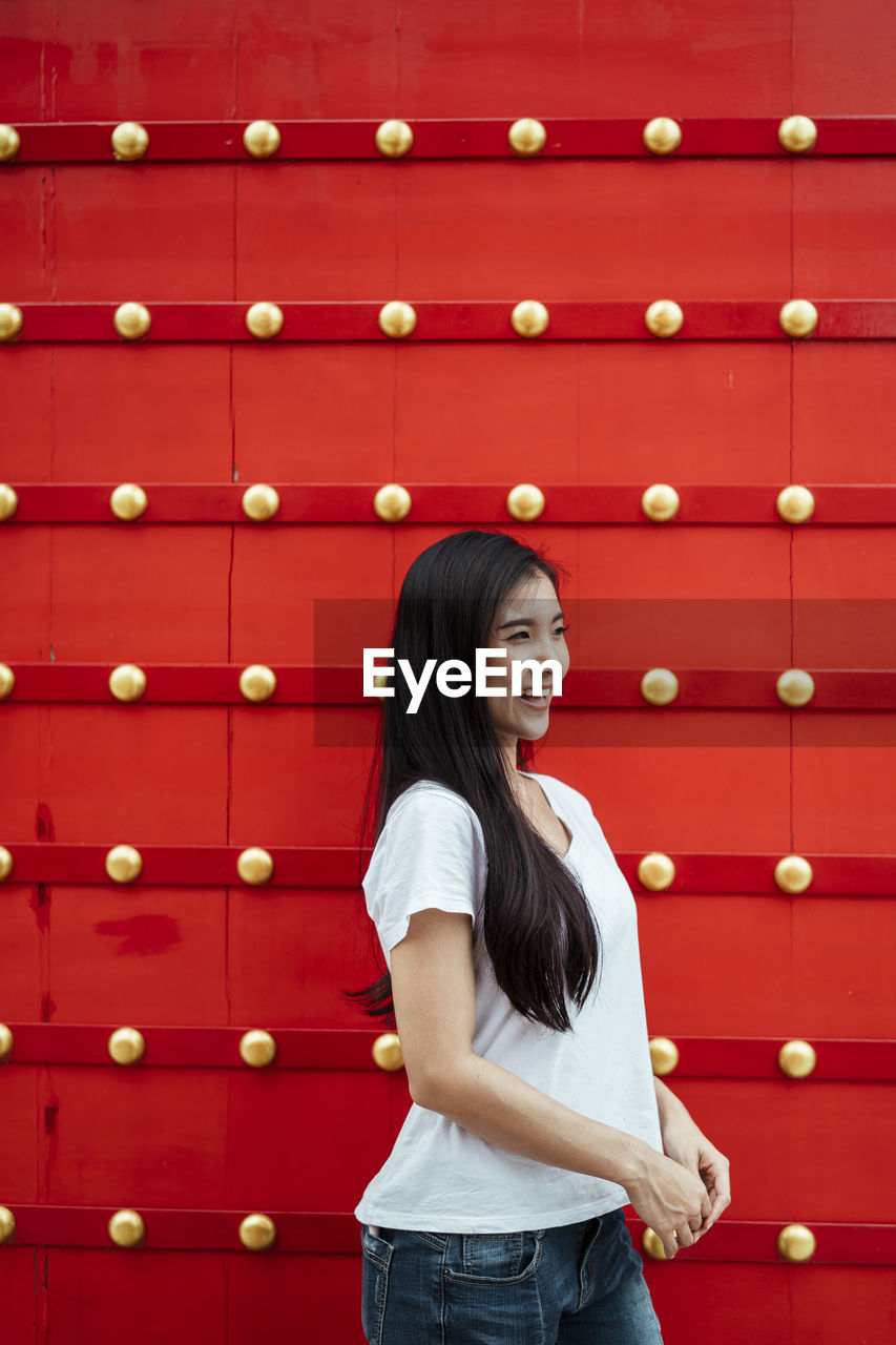 FULL LENGTH OF A YOUNG WOMAN STANDING AGAINST RED WALL