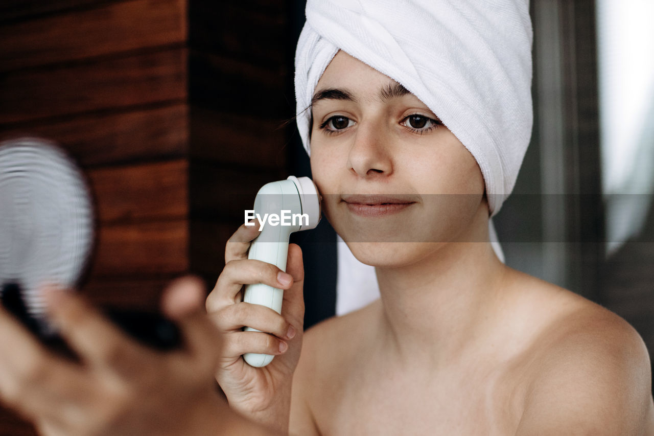 Young girl with towel on head makes facial massage with wireless electric massager