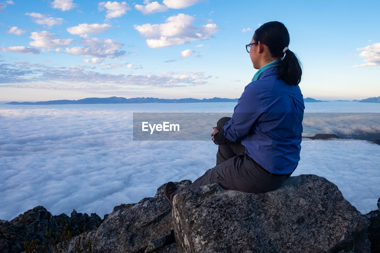 Woman in blue hiking shirt sitting on rock looking at clouds below 