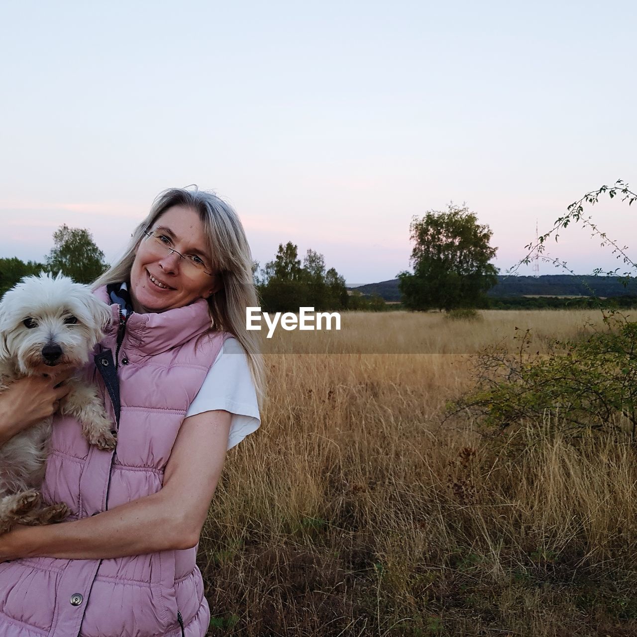 Portrait of smiling mature woman with dog standing on field against sky during sunset