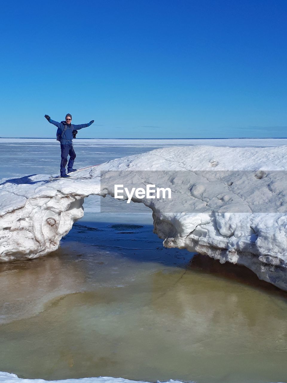 Man with arms outstretched standing on glacier in sea against blue sky