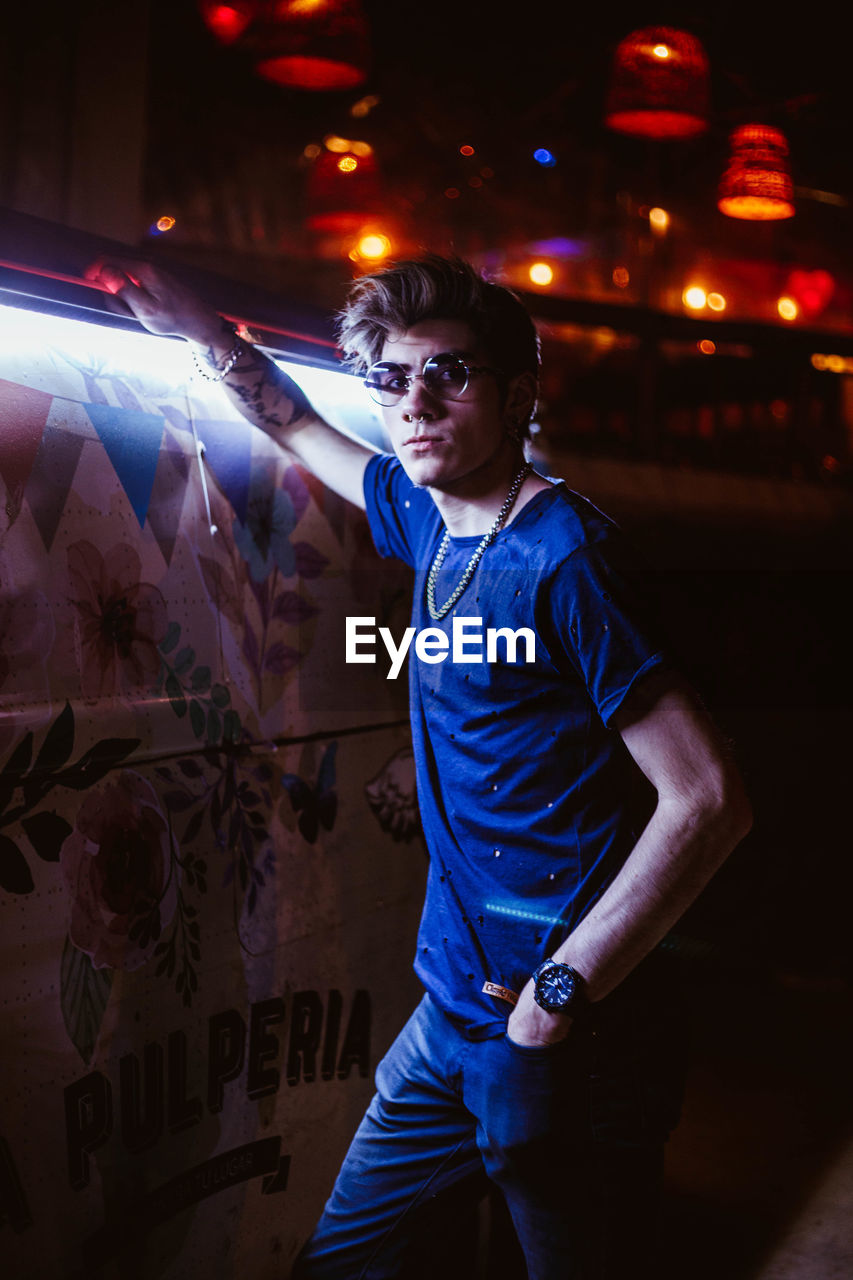 FULL LENGTH PORTRAIT OF YOUNG MAN STANDING AGAINST ILLUMINATED WALL