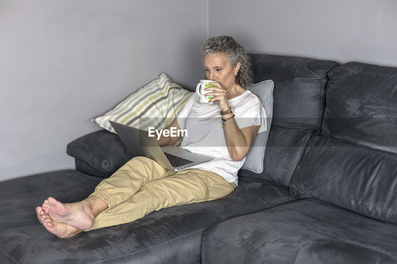 A woman on the sofa using a laptop for remote work and drinking.