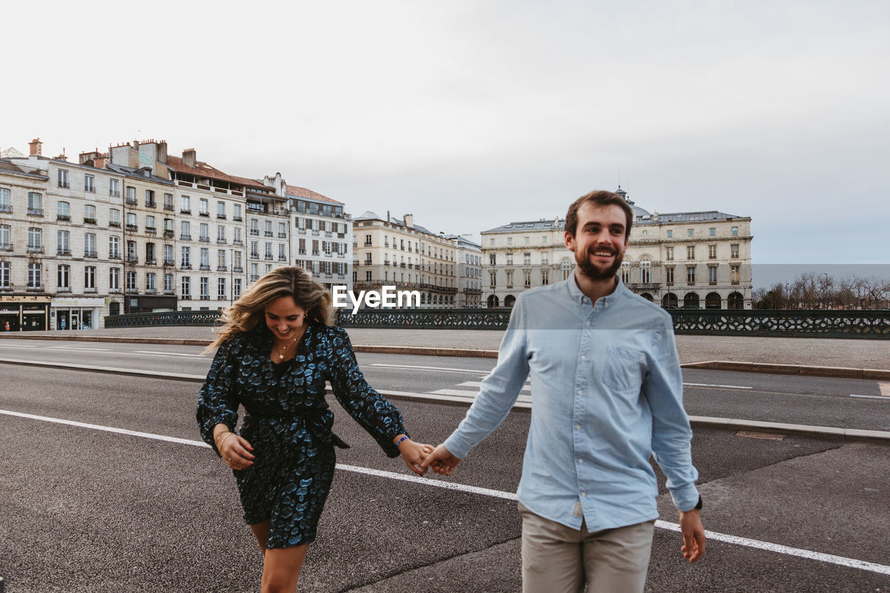 Happy young romantic couple in stylish clothes laughing and holding hands while crossing bridge with historic buildings in background during city tour in bayonne in france