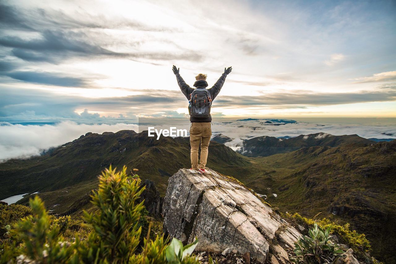 Full length rear view of man with arms raised standing on rock at mountain against sky