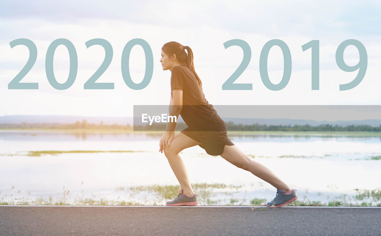 Digital composite image of young woman stretching on road by 2020 and 2019 against lake and sky