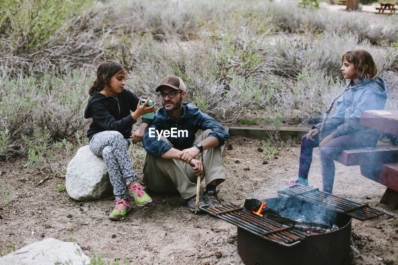 Girl looking at sister showing smart phone to father while sitting at campsite