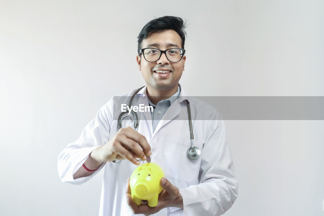 Portrait of smiling doctor holding piggy bank against white background