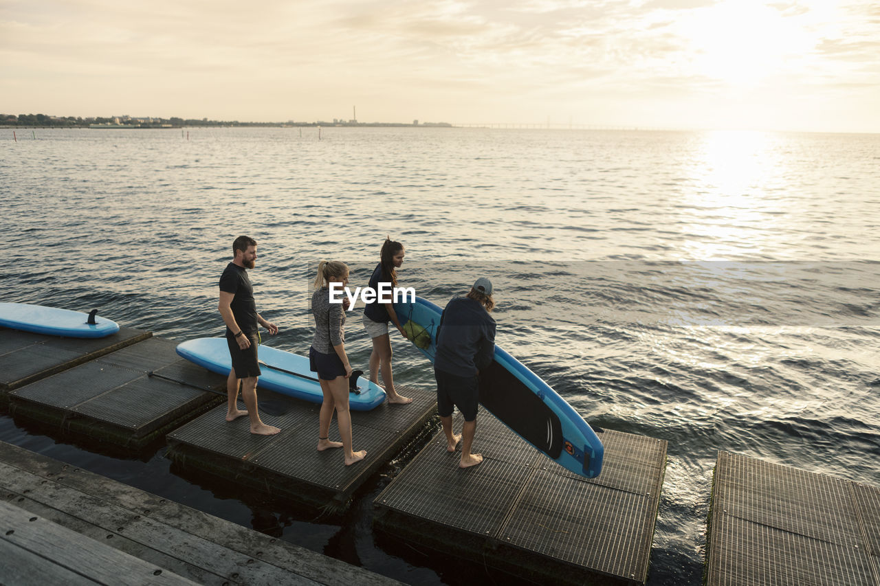 Male and female friends carrying paddleboard while standing on pier by sea