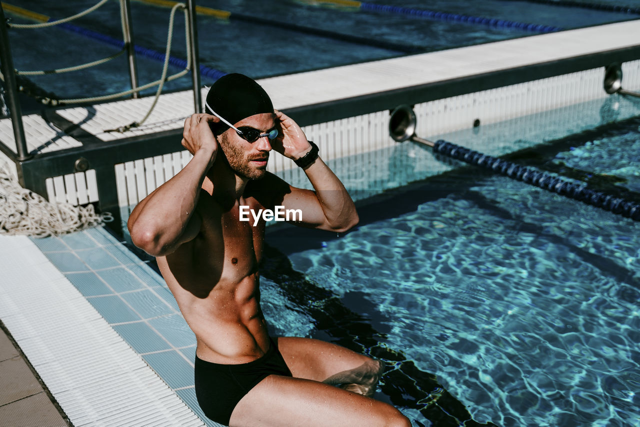 Male swimmer adjusting cap while sitting at poolside