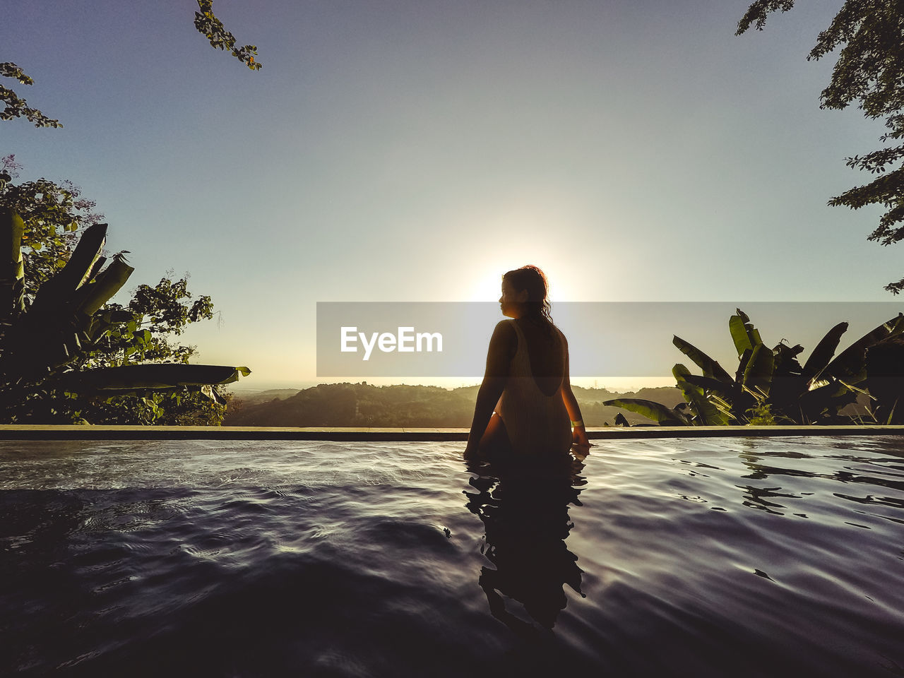 Rear view of standing by swimming pool against sky during sunset