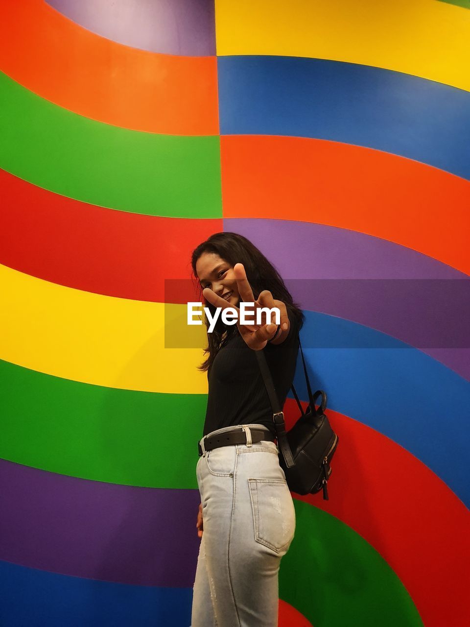 Portrait of young woman showing peace sign while standing against colorful wall