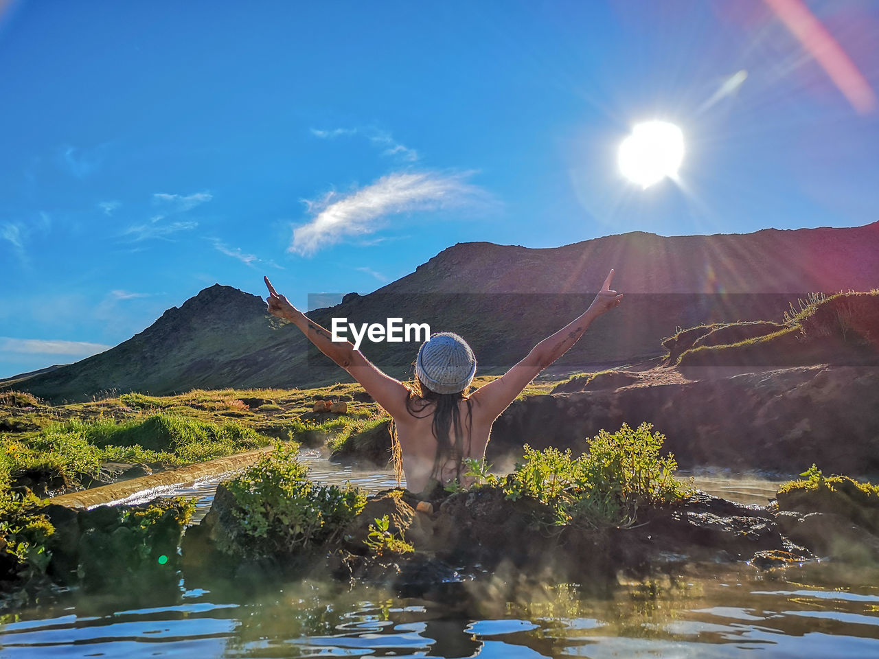 Rear view of woman with arms raised at hot spring against mountain
