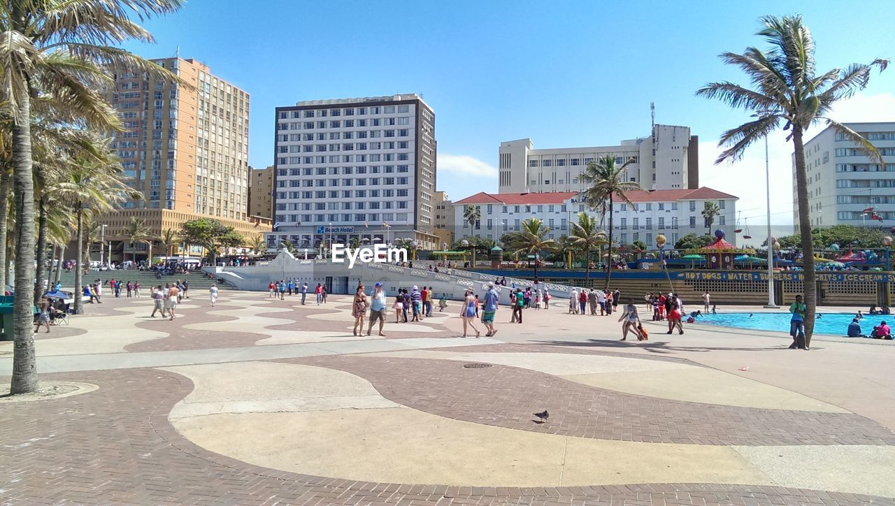 town square, plaza, walkway, city, architecture, building exterior, built structure, boardwalk, downtown, crowd, group of people, large group of people, tree, public space, nature, neighbourhood, sky, palm tree, travel destinations, building, day, tropical climate, city life, vacation, plant, street, office building exterior, shopping mall, urban area, sunny, sunlight, beach, outdoors, skyscraper, travel, sports, clear sky, park