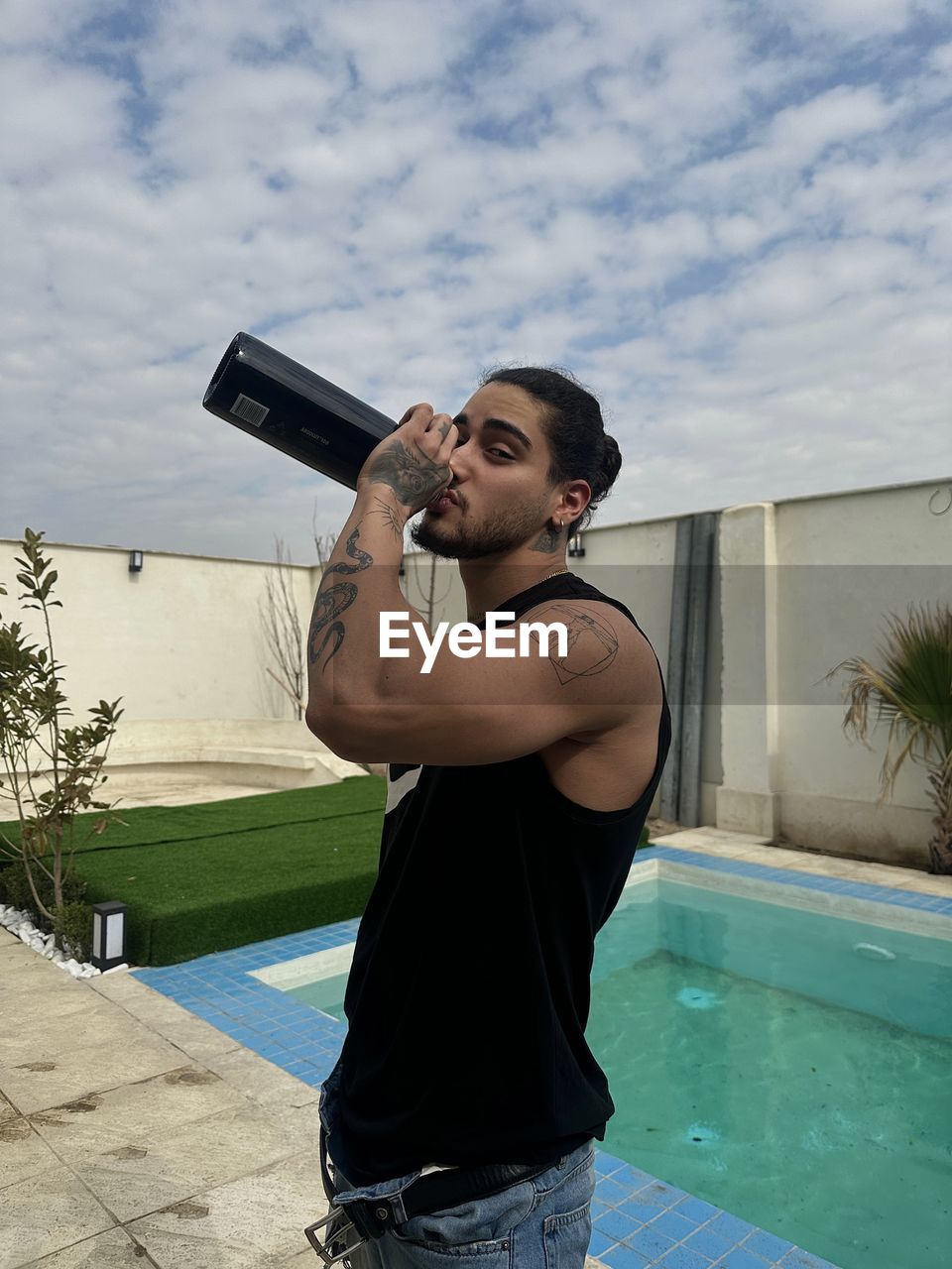 Side view of young man drinking water while standing against swimming pool