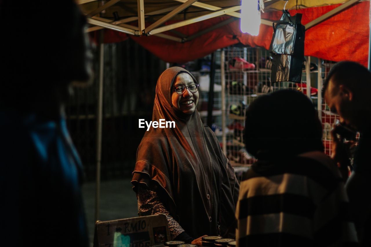 Cheerful female vendor with customers at illuminated market stall during night