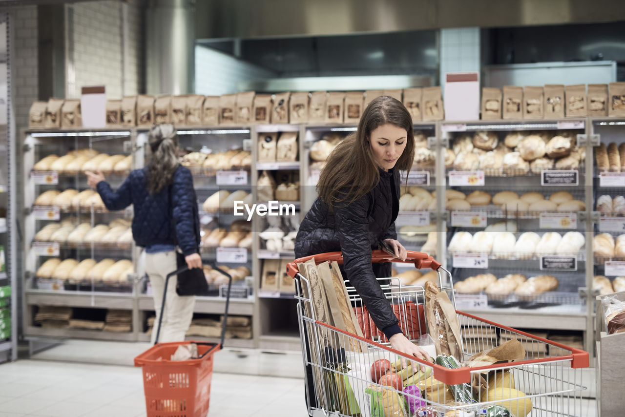 Woman keeping bananas in shopping cart against rack at supermarket