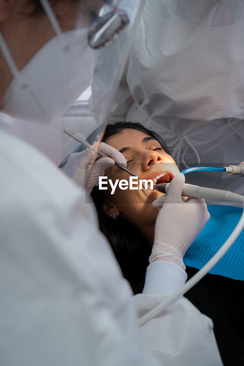 Crop professional dentist in uniform with medical mask drilling tooth of calm woman with help of assistant
