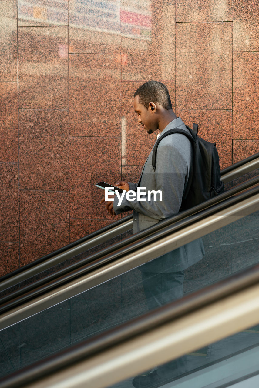 Side view of african american male worker listening to music in tws earbuds and browsing cellphone on escalator