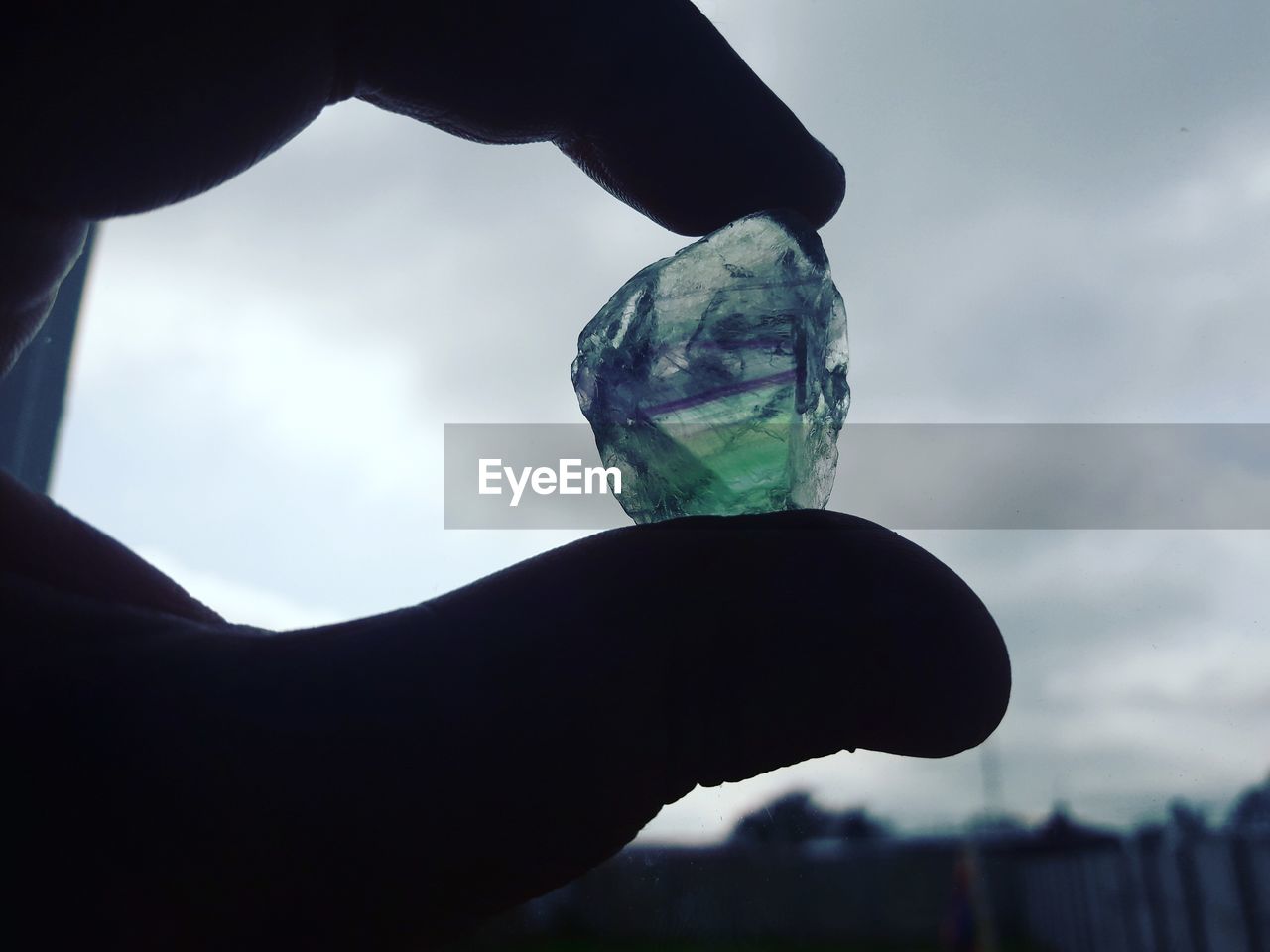 Close-up of cropped silhouette hand holding gemstone against cloudy sky