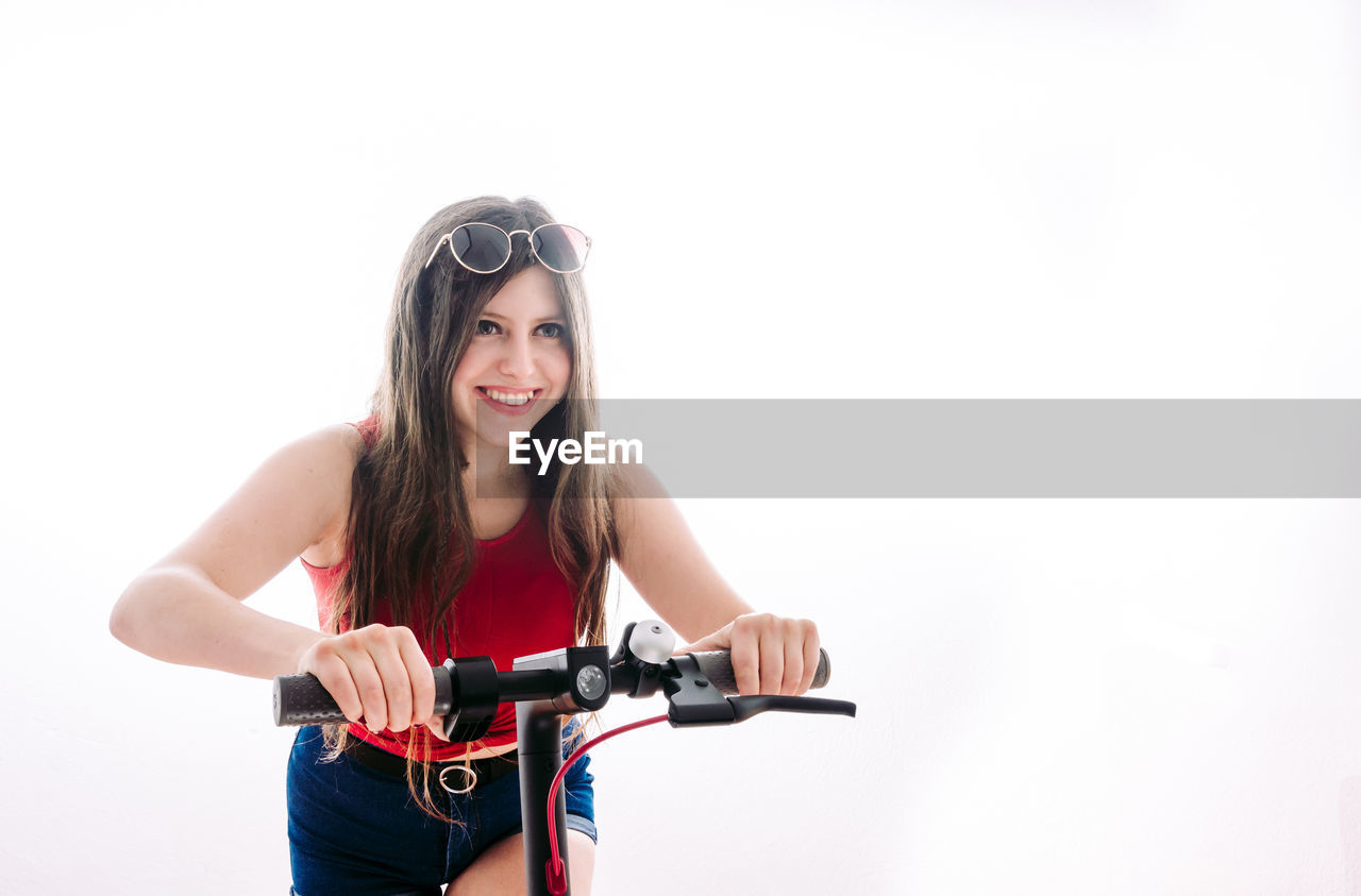 PORTRAIT OF HAPPY YOUNG WOMAN RIDING BICYCLE AGAINST WHITE BACKGROUND