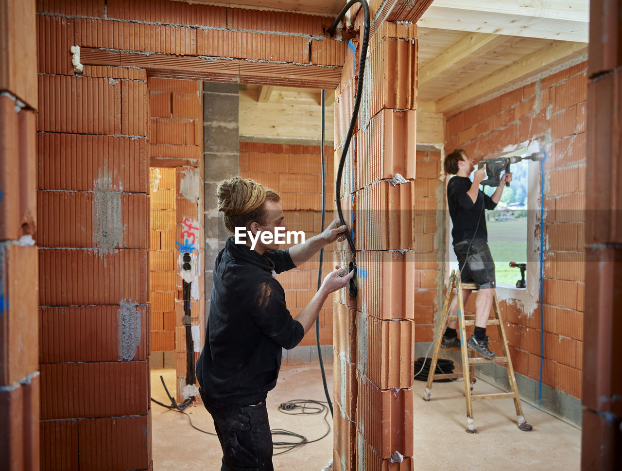 Male electricians installing cables in brick wall while colleague working in background at construction site