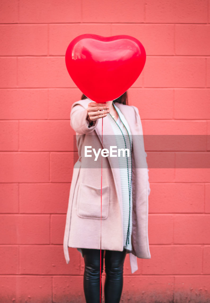 Woman holding heart shape balloon against wall