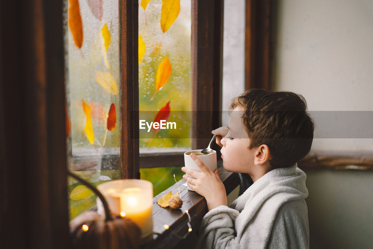 Boy wrapped in a blanket drink hot tea and looks out the open window at the wonderful autumn nature