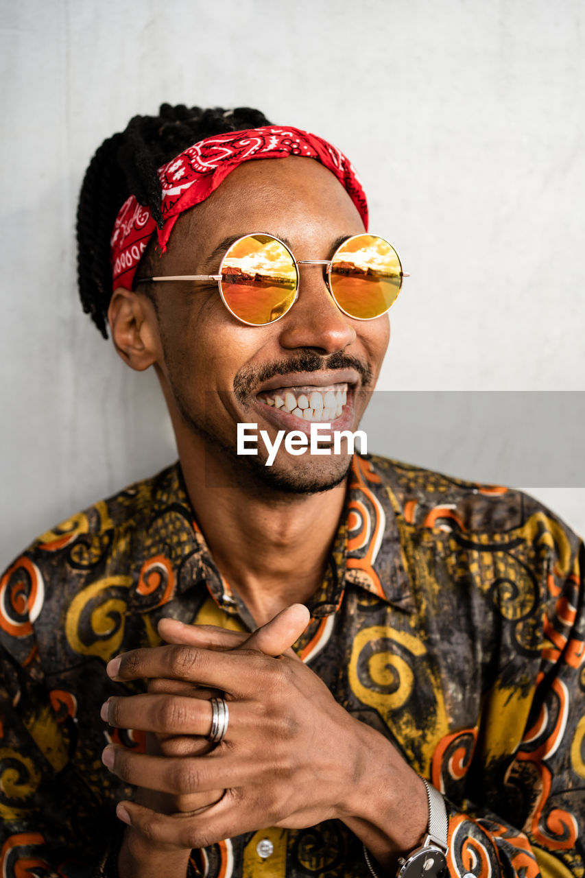 Happy african american guy with dreadlocks wearing stylish round sunglasses with red headband and colorful shirt smiling away