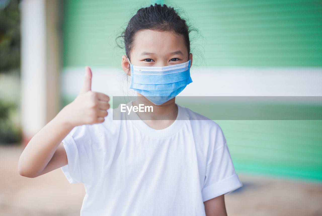 Portrait of girl wearing mask showing thumbs up white while standing outdoors