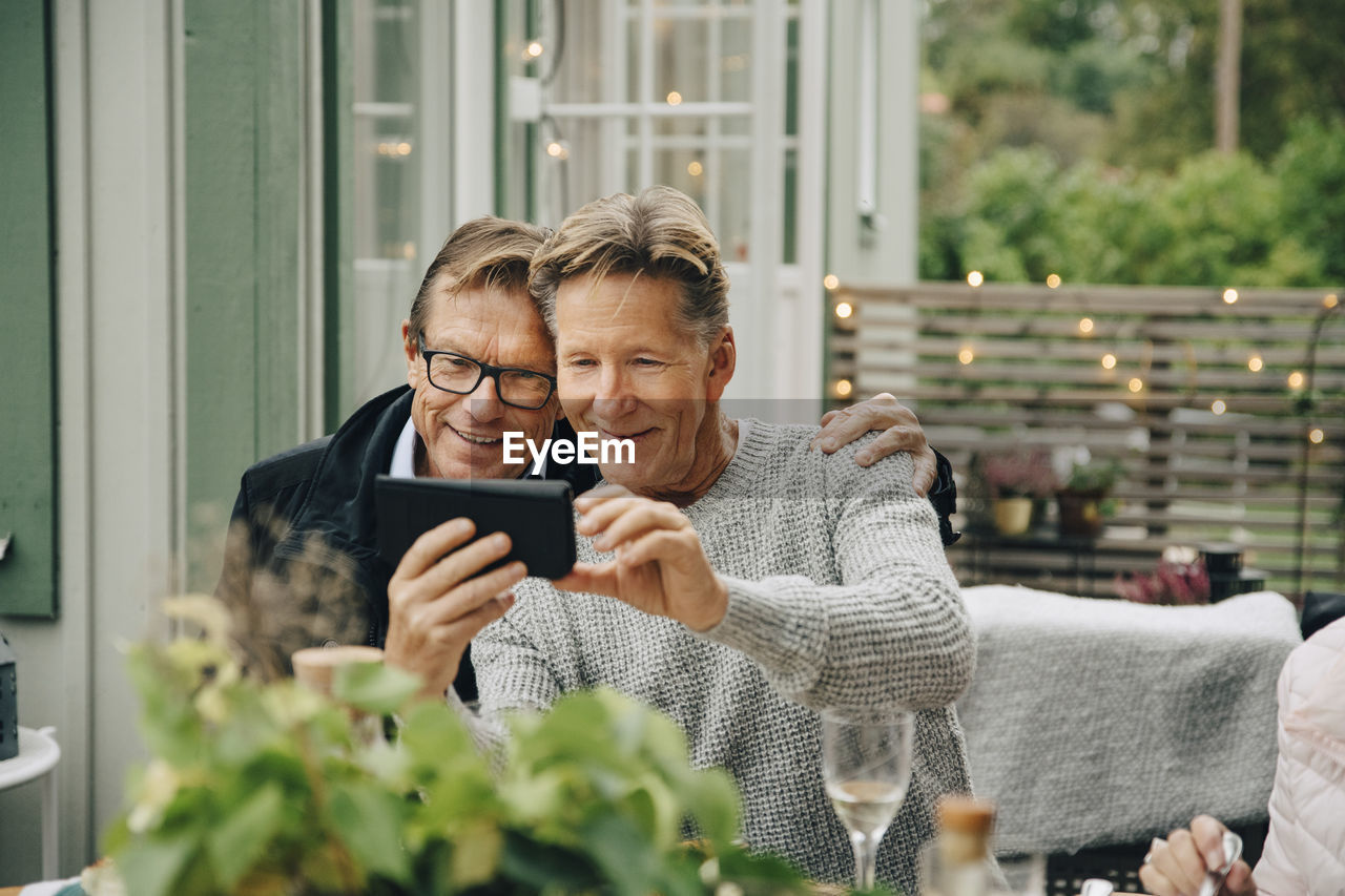 Smiling man showing smart phone with friend while sitting at back yard during party