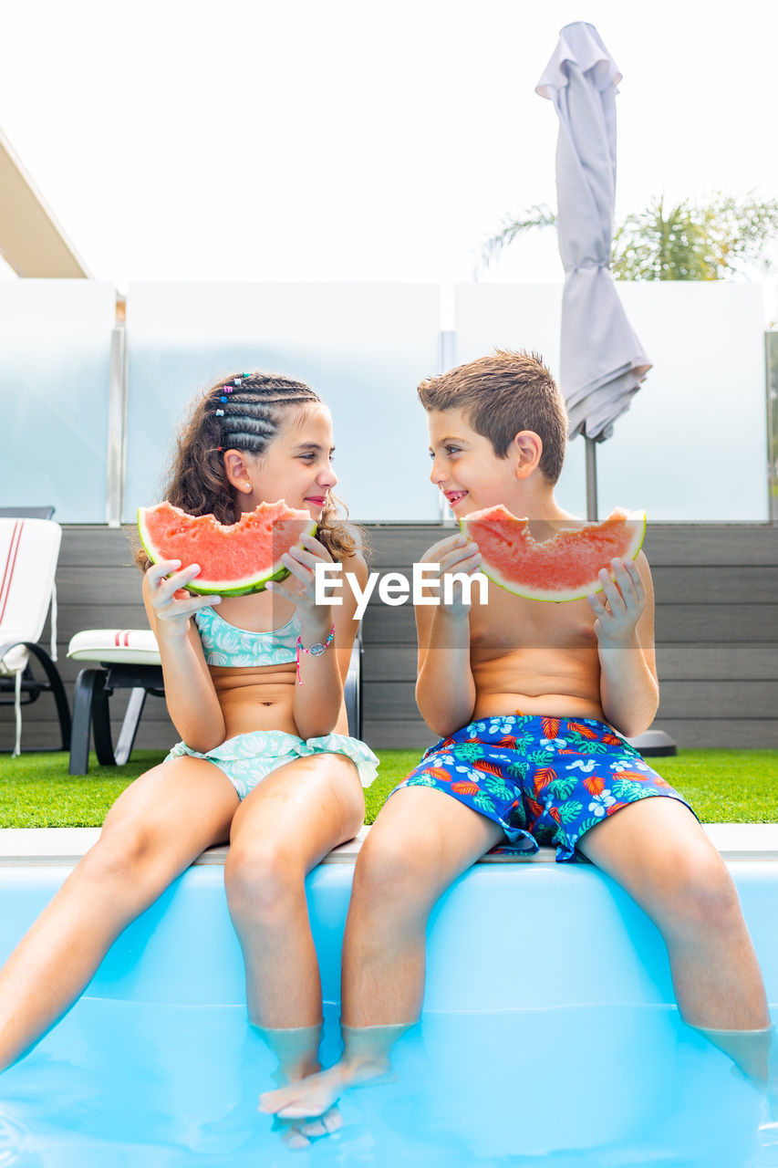 Two funny kids eating watermelon at the the edge of the pool on a summer day