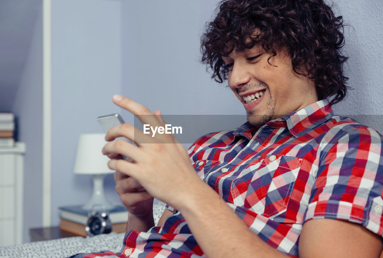 Close-up of smiling man using mobile phone while sitting on bed at home