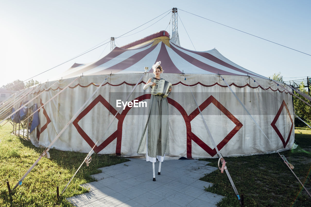 Female circus performer playing accordion while standing on stilts in front of circus tent