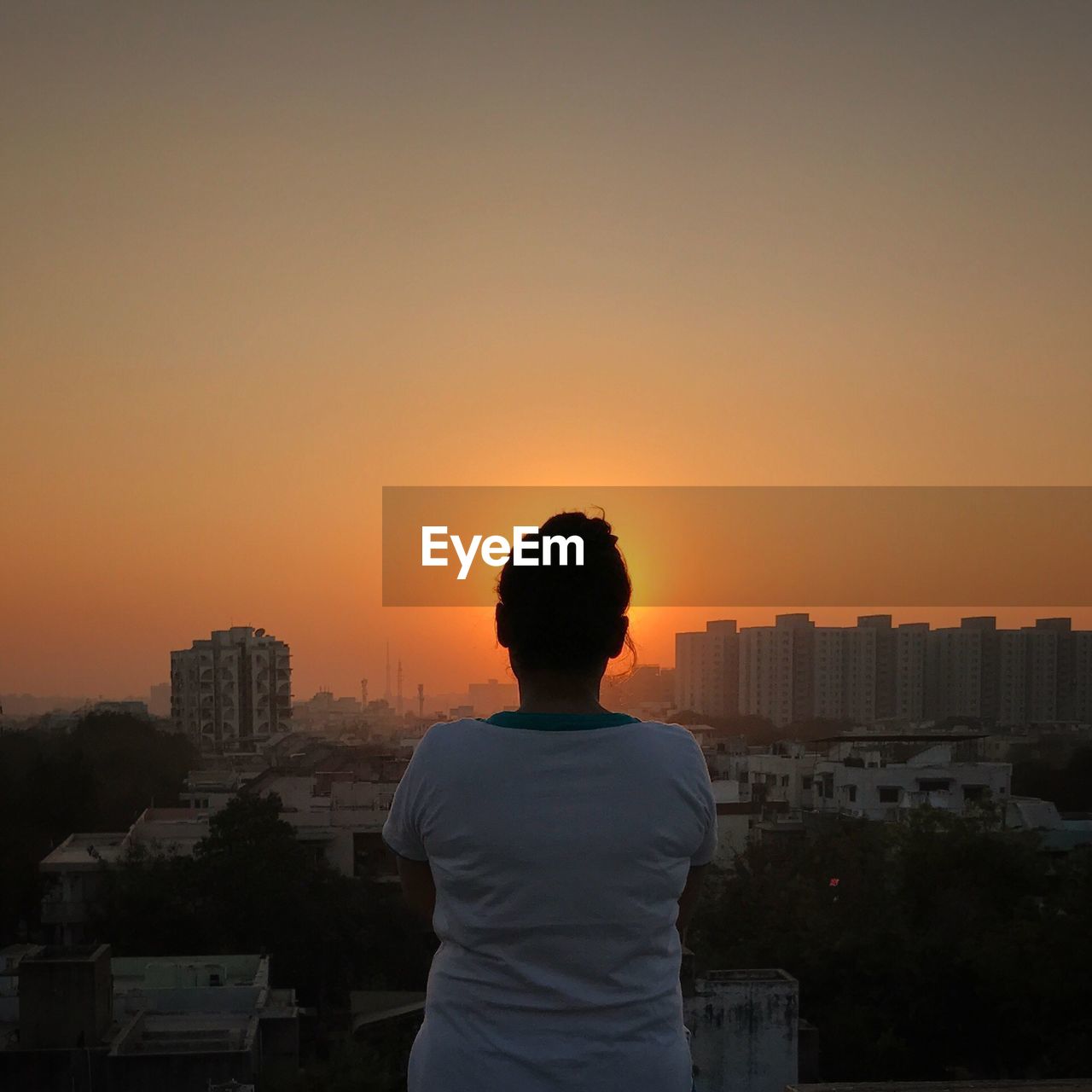 REAR VIEW OF MAN LOOKING AT CITYSCAPE AGAINST SKY DURING SUNSET