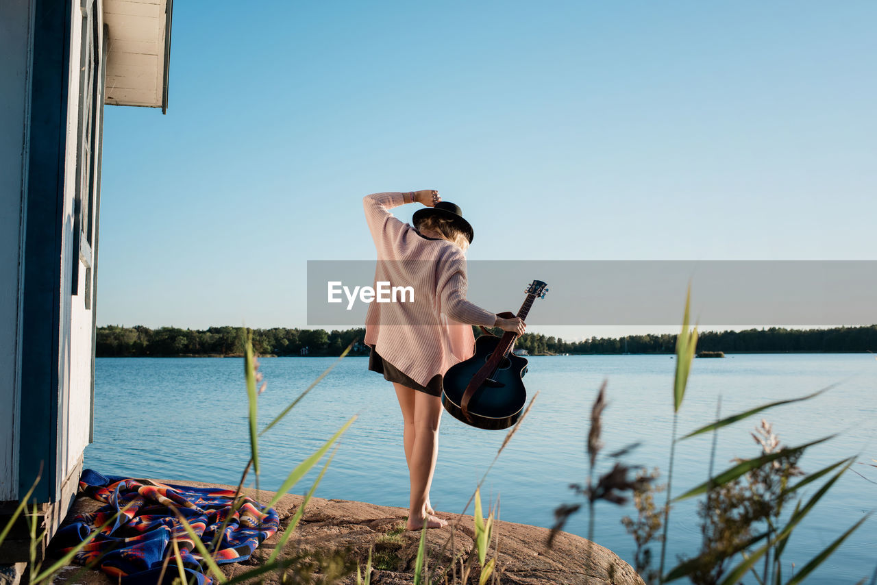 Woman walking on a rock at the beach holding her guitar in the sun