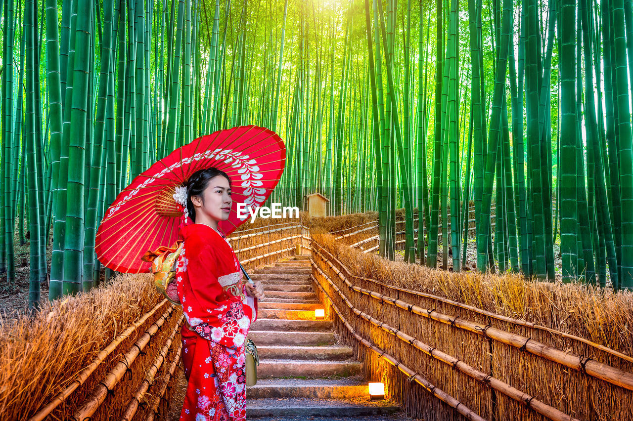 Mid adult woman holding red umbrella on steps amidst bamboo forest