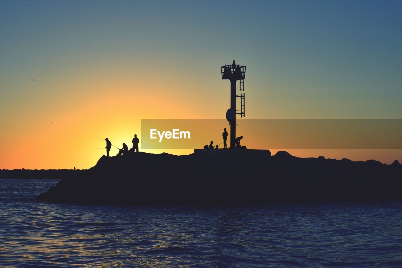 Silhouette men on rock by sea against sky during sunset