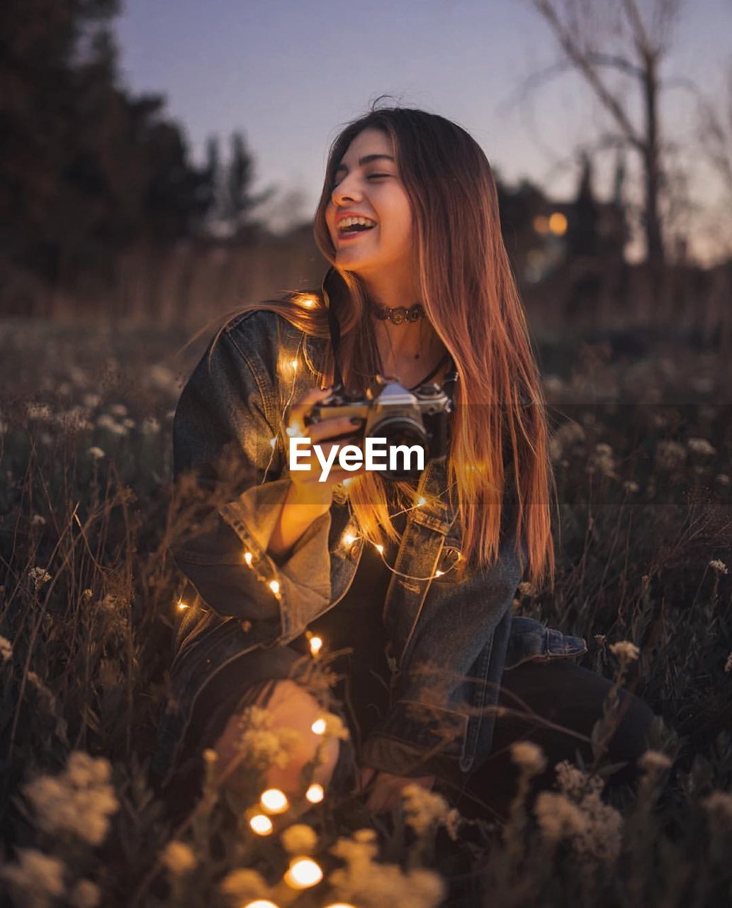 Young woman smiling while holding camera and illuminated string lights while sitting amidst plants on land 