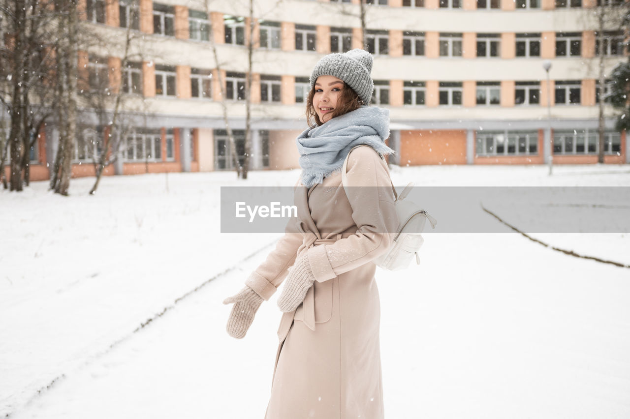 A cute girl under the snow with a backpack goes to the building and smiles