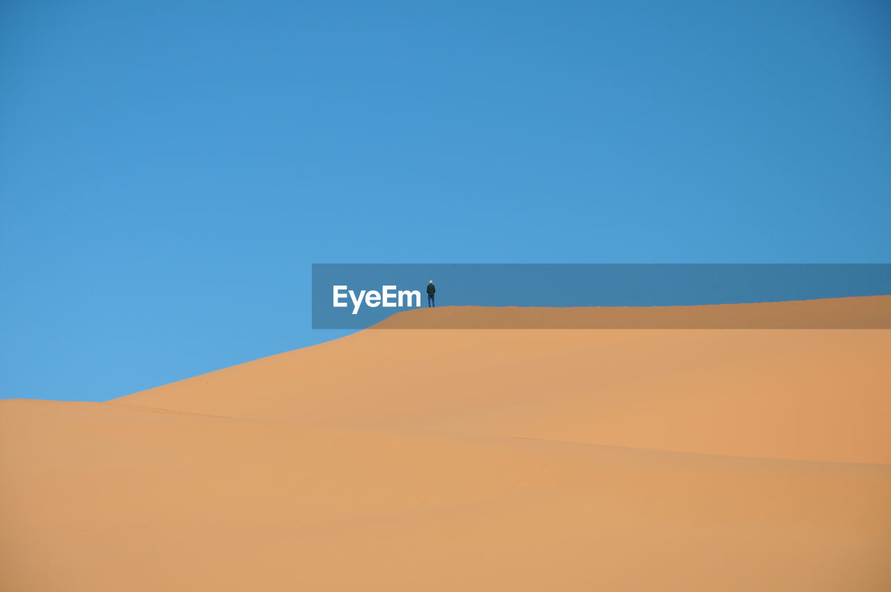 Distant view of person standing on sand dune against clear blue sky