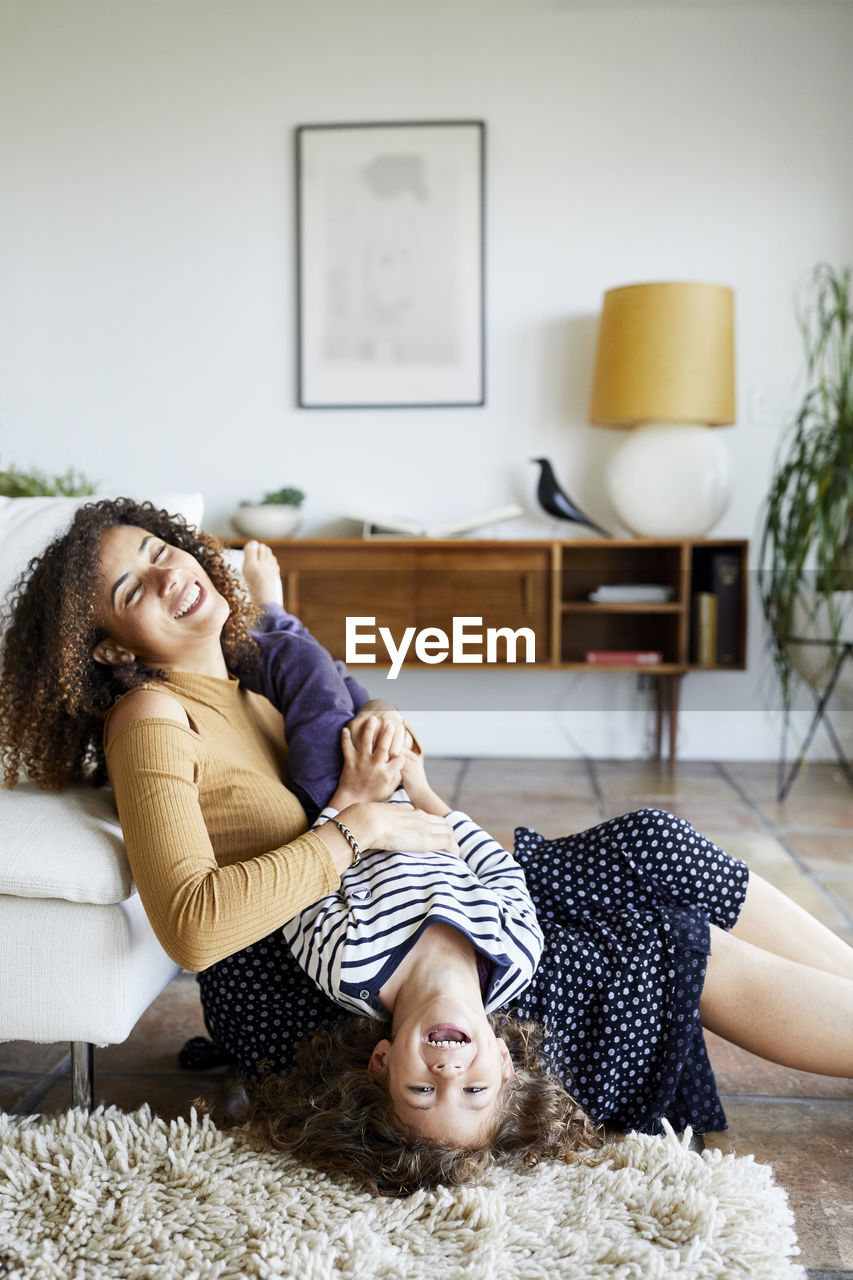 Happy mother holding daughter upside down while sitting on floor by sofa at home