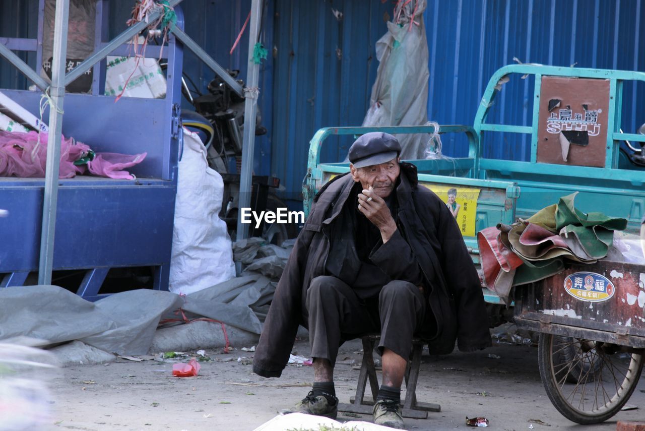 Homeless man smoking cigarette while sitting on stool in city