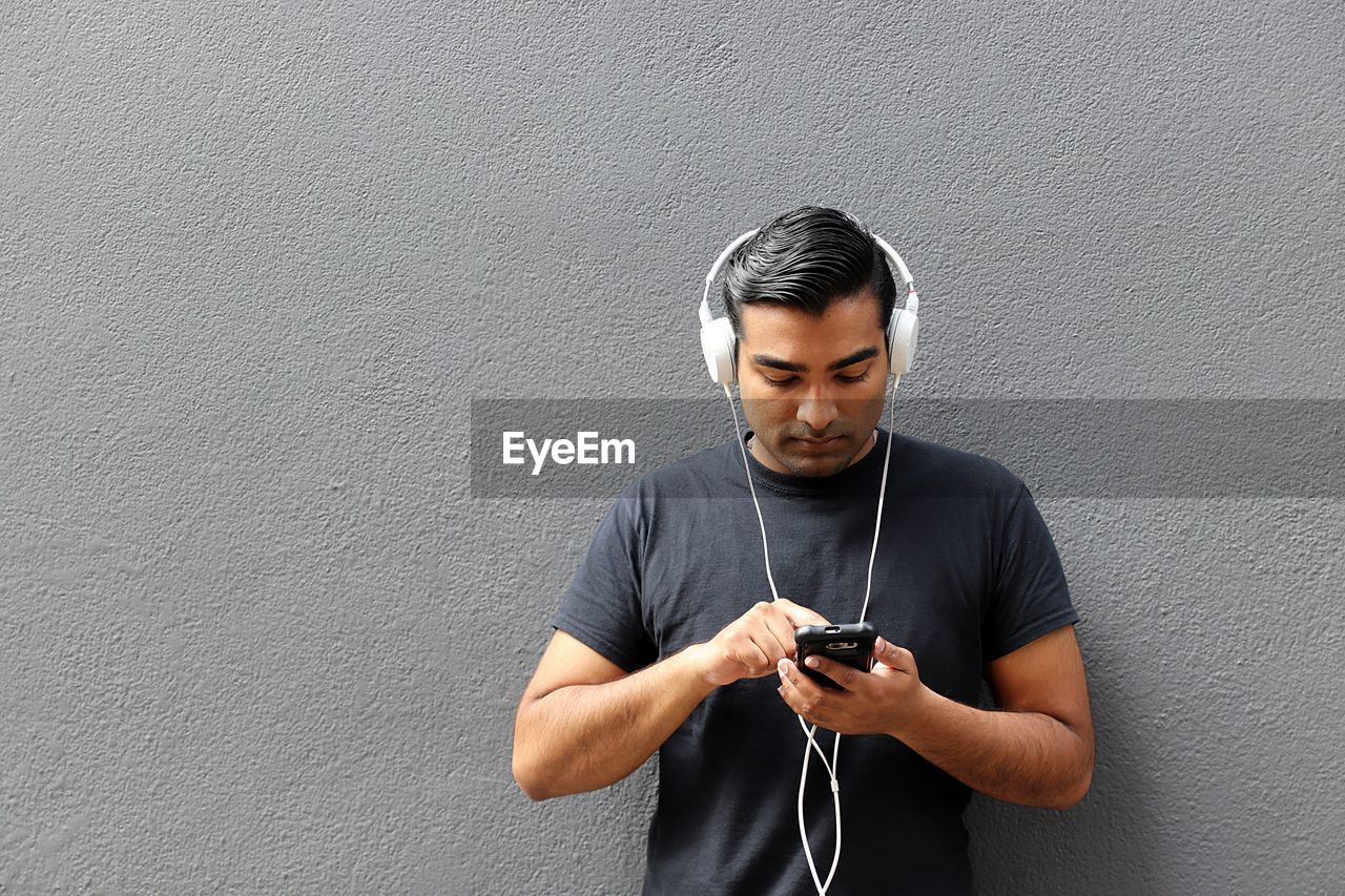 Young man using mobile phone while listening music against wall