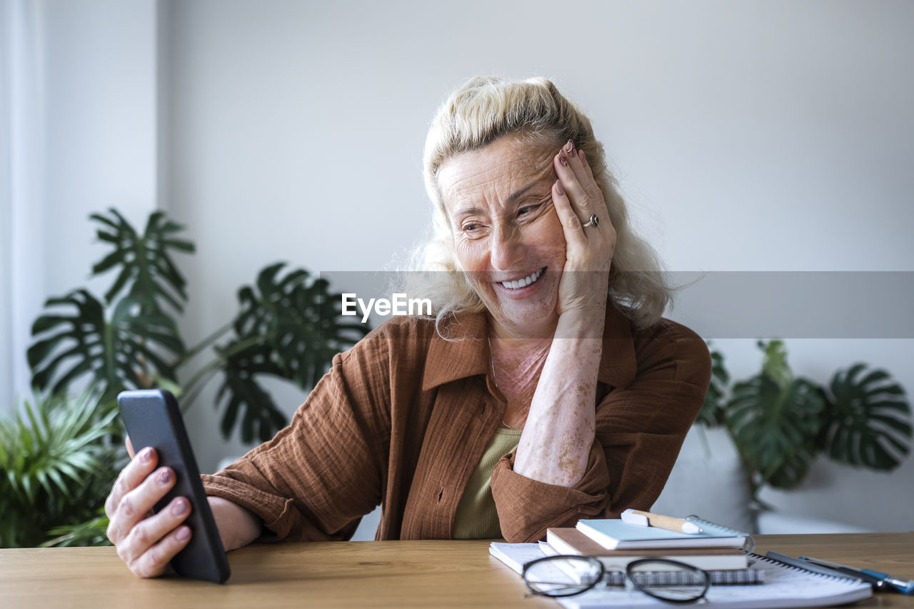 Smiling freelancer watching mobile phone at desk in home office