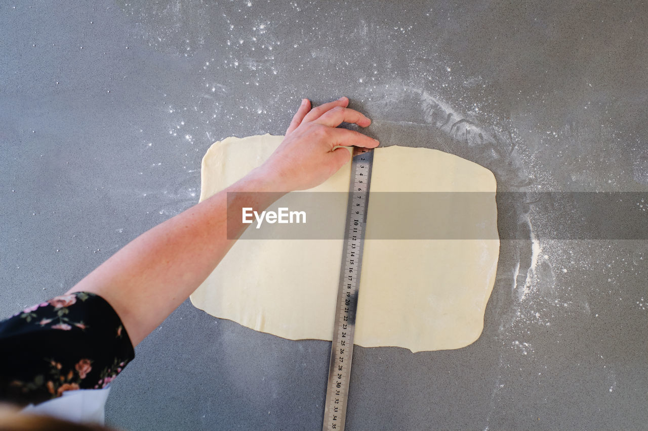 From above of crop unrecognizable female measuring handmade rolled dough placed on gray table in daytime